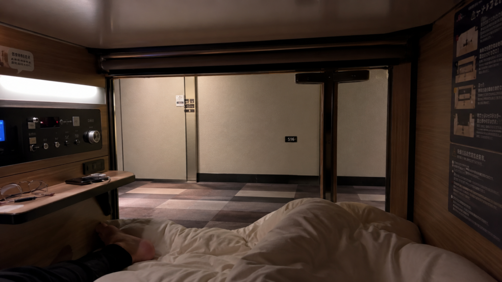 Photorealistic 16:9 photo taken from inside a capsule hotel pod looking out, showing the compact interior with a folded duvet, small shelf with phone and glasses, control panel with dimmed light settings, view through the open blind to a quiet corridor, warm natural tones, authentic traveller perspective, realistic phone-camera quality, no filters.